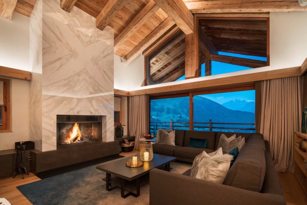 A cozy living room with a modern fireplace, brown sectional sofa, and large windows showing a mountain view at dusk. Exposed wooden beams and soft lighting create a warm, inviting atmosphere. David Pearson Travel