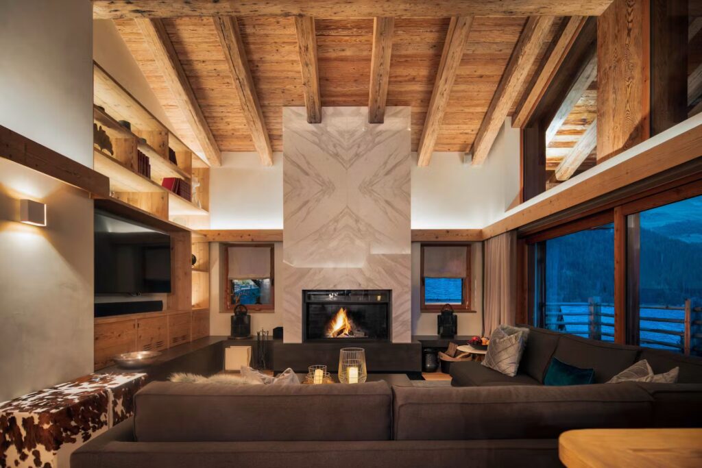 A cozy, modern living room with exposed wooden beams, a stone fireplace with a lit fire, large windows showing a mountain view, and comfortable sofas arranged around the hearth. David Pearson Travel