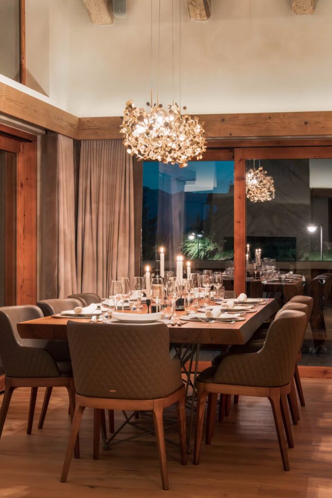 A warmly lit dining room with a wooden table set for a meal, surrounded by cushioned chairs. Elegant chandeliers hang above, with candles and glassware adding to the cozy, inviting ambiance. Large windows reflect the scene. David Pearson Travel