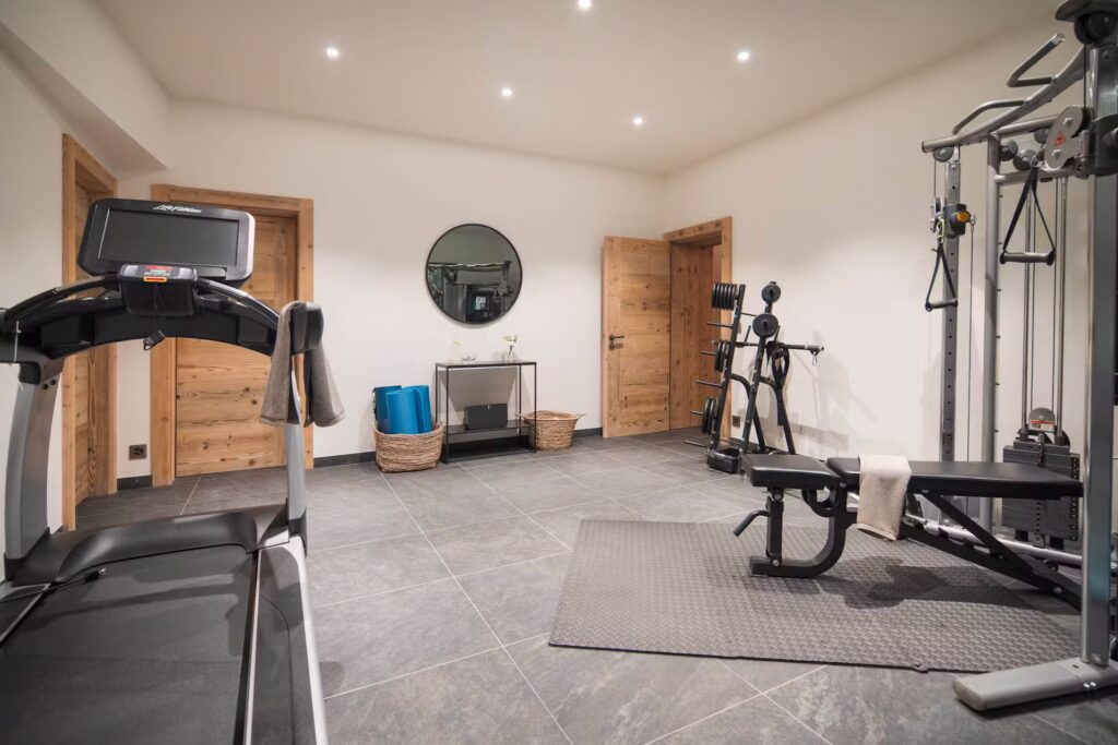 Modern home gym with treadmill, weight bench, multi-gym machine, and exercise mats. Wooden doors, round wall mirror, baskets, and minimal decor. Gray tiled floor with towels on gym equipment. David Pearson Travel
