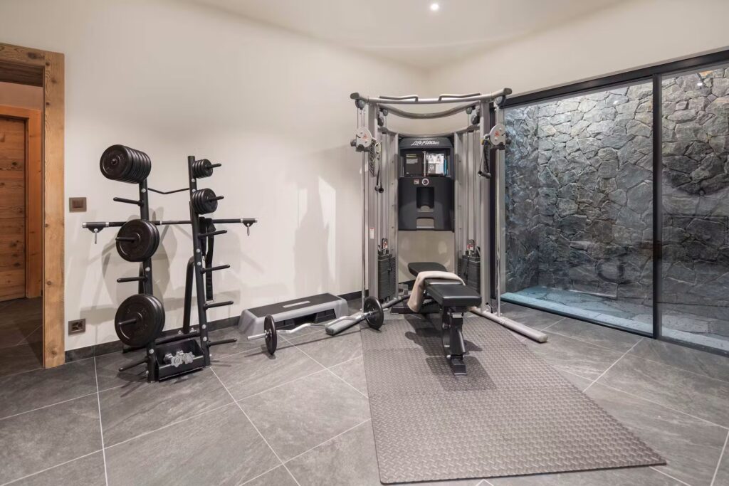 A modern home gym with weight plates on a rack, a bench press, a multi-gym machine, a barbell, and a treadmill. The room has slate flooring and a large window showing a stone wall outside. David Pearson Travel