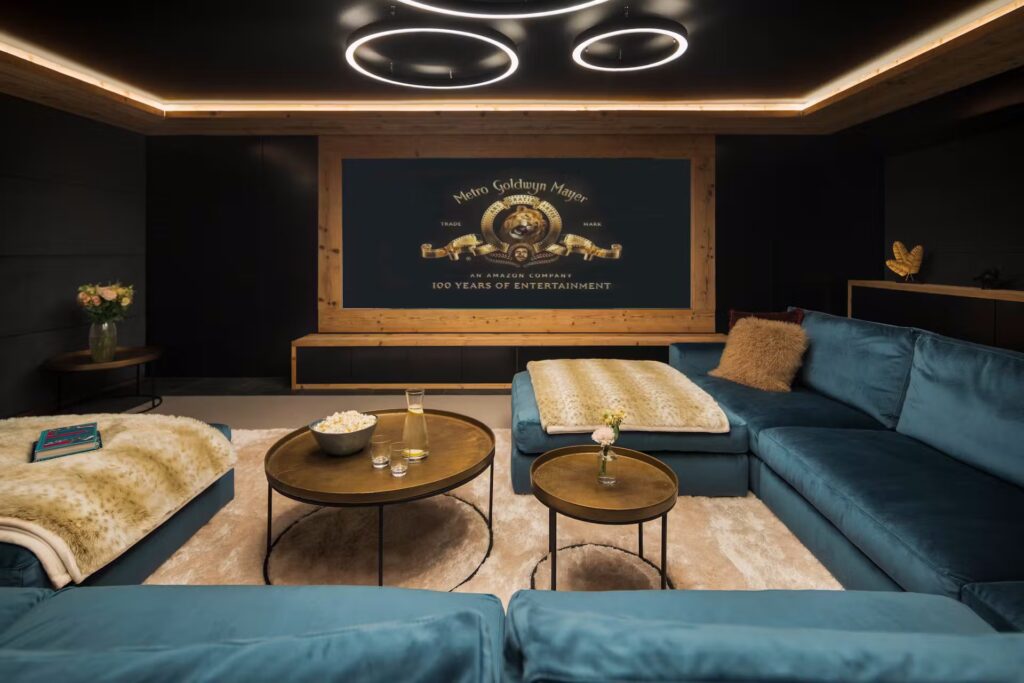 A modern home theater with blue velvet sectional sofas, round wooden tables with drinks and popcorn, cozy blankets, and a large MGM movie screen framed in wood. Circular ceiling lights illuminate the stylish space. David Pearson Travel