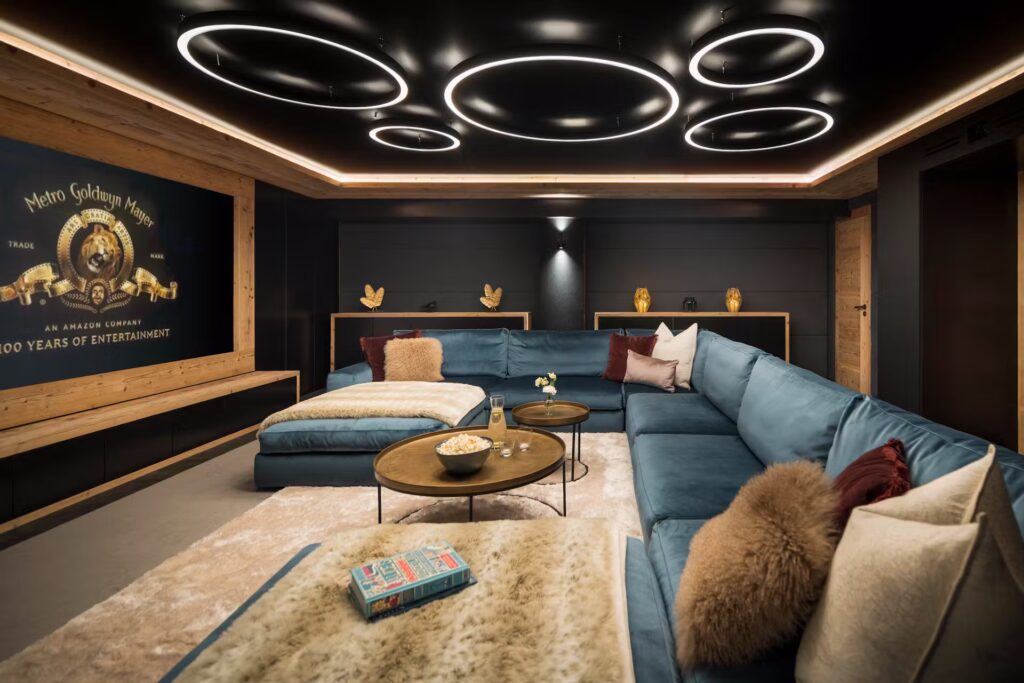 A modern home theater with a large blue sectional sofa, round coffee tables, plush pillows, and a beige rug. The wall features a Metro-Goldwyn-Mayer logo, and the ceiling has circular LED lights. David Pearson Travel