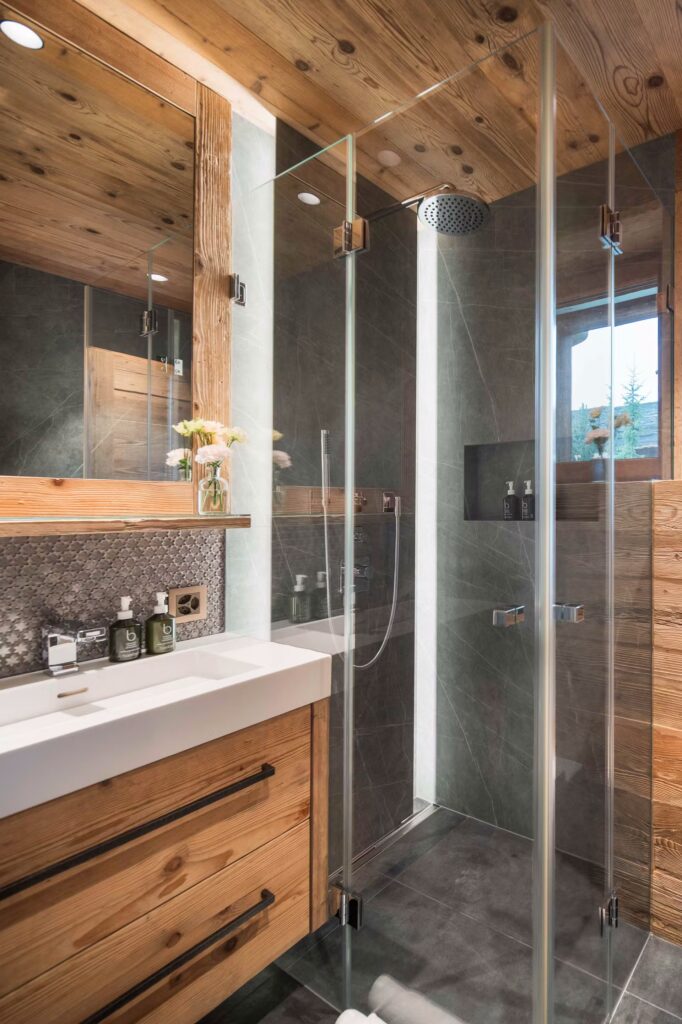 A modern bathroom with wood accents, a glass shower enclosure, rainfall showerhead, grey tile walls and floors, a white sink, a large mirror, and toiletries neatly arranged on the sink. David Pearson Travel