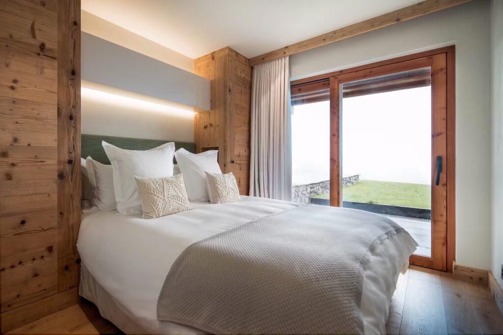 A cozy bedroom with a double bed, white bedding, and decorative pillows. Wooden walls and flooring add warmth. Large glass doors open to a patio with grass and a stone wall, letting in natural light. David Pearson Travel