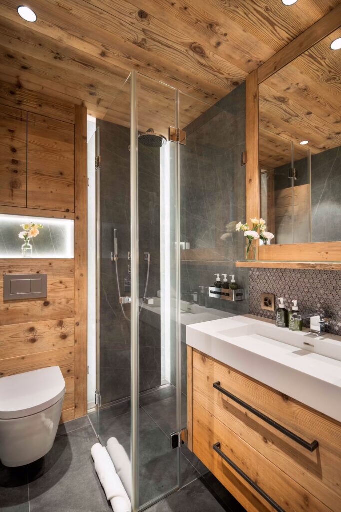 A modern bathroom with wood-paneled walls and ceiling, a glass shower enclosure, a white toilet, a rectangular sink with wood cabinetry, a large mirror, hexagonal tile backsplash, and fresh flowers on the counter. David Pearson Travel