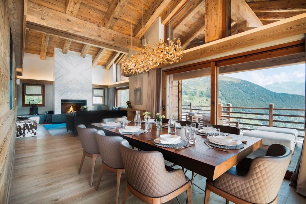 A cozy, modern dining room with a wooden table set for six, elegant upholstered chairs, a unique chandelier, large windows, and mountain views. Exposed wooden beams and a fireplace add warmth to the space. David Pearson Travel