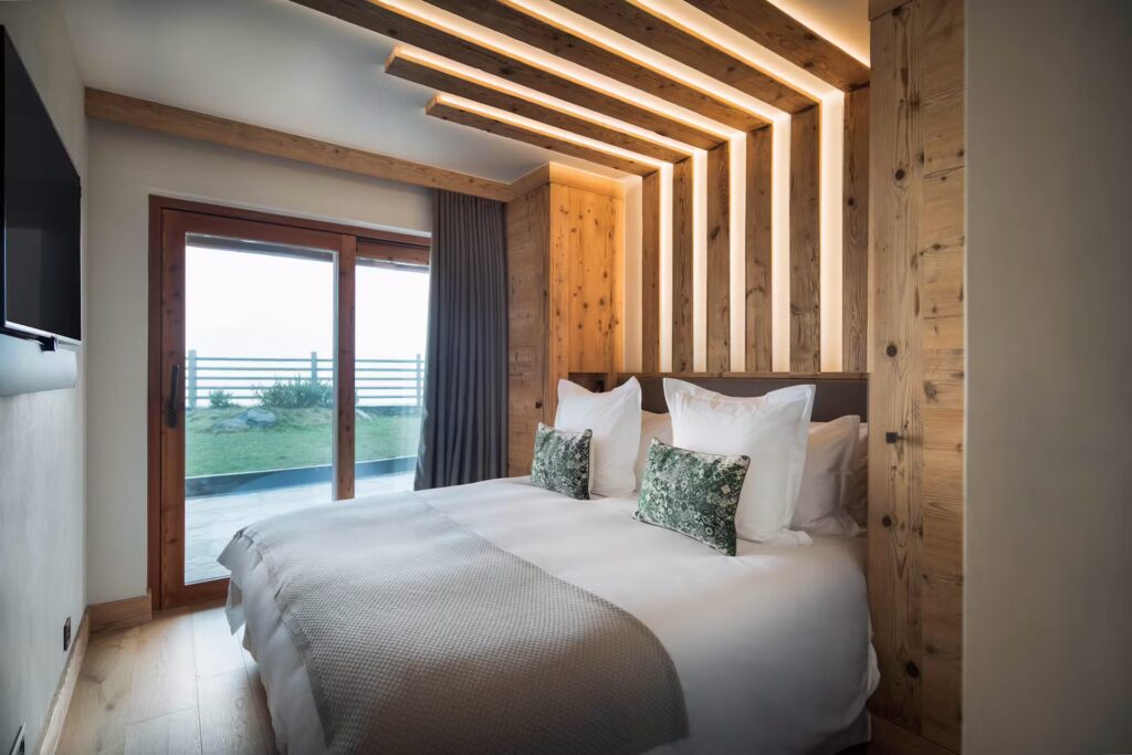 A modern bedroom with a large bed, white bedding, green accent pillows, wood paneling, warm lighting, a wall-mounted TV, and sliding glass doors leading to a patio with a grassy area outside. David Pearson Travel