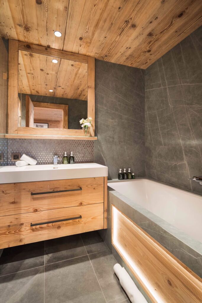 A modern bathroom with wood panel ceiling and vanity, grey tiled walls and floor, a large mirror, a bathtub with built-in lighting, and toiletries arranged neatly on the sink and tub. David Pearson Travel