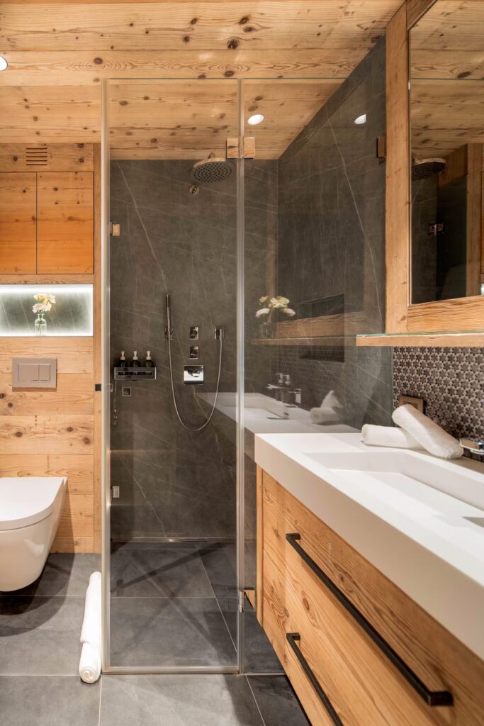 Modern bathroom with wood paneling, a glass-enclosed shower, dual sinks with a large mirror, floating toilet, gray tile floor, and rolled white towels on the counter and floor. Subtle lighting adds warmth to the space. David Pearson Travel