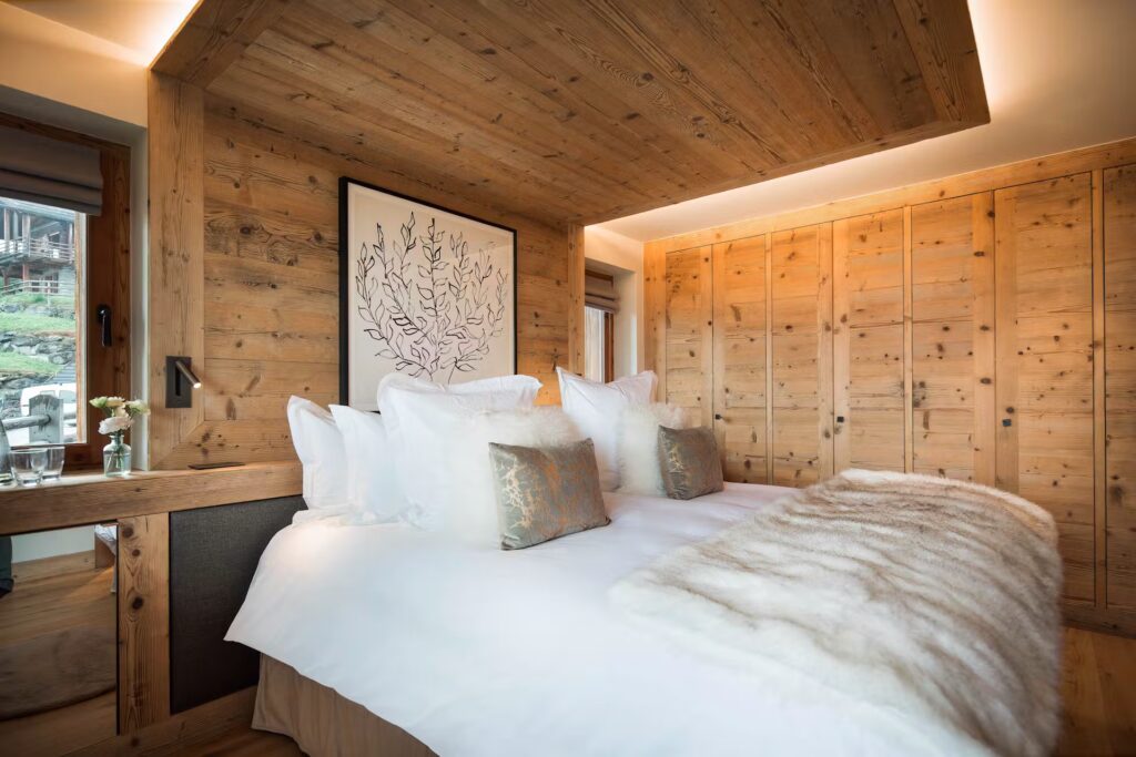 A cozy, modern bedroom featuring wood-paneled walls and ceiling, a large bed with white linens and fluffy pillows, fur throw, built-in wooden wardrobes, and decorative artwork above the headboard. David Pearson Travel