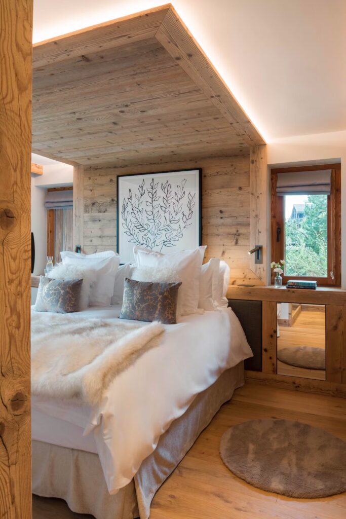 A cozy bedroom with a wooden accent wall and ceiling, a large bed with white linens and fur throw, decorative pillows, a round rug, a desk with a mirror, and a window letting in natural light. David Pearson Travel