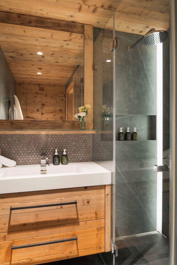 Modern bathroom with wood accents, a white sink, and a glass shower. A large mirror, flowers in a vase, and bottles of toiletries sit on the counter and in the shower niche. The lighting is warm and inviting. David Pearson Travel