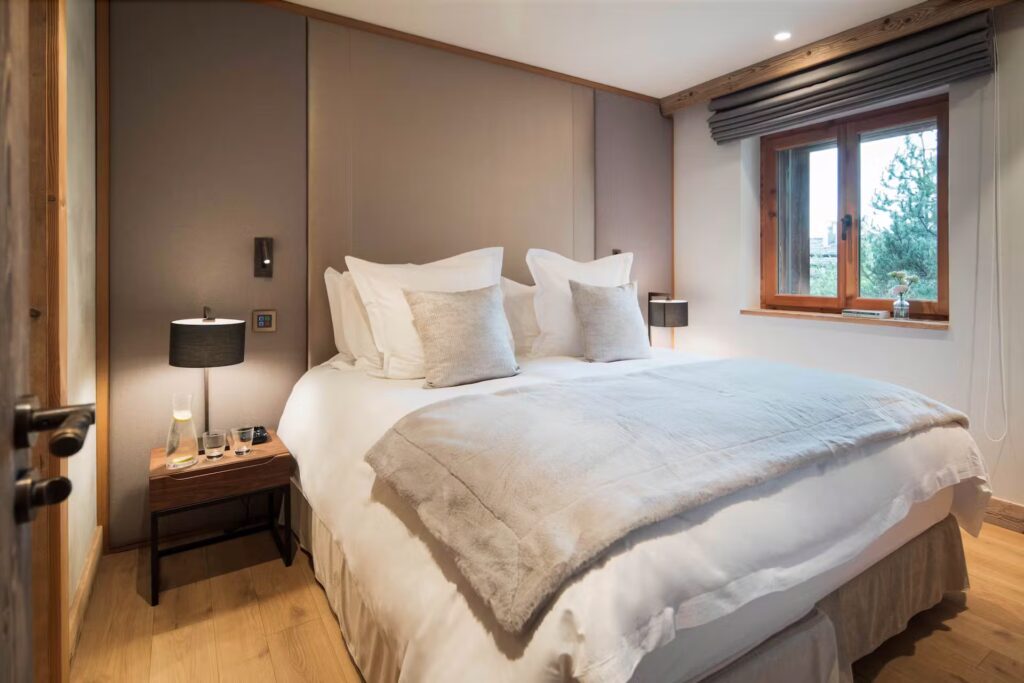 A cozy bedroom with a large bed, white bedding, beige throw and pillows, wooden side table with a lamp and glasses, a window with gray blinds, and soft lighting creating a warm atmosphere. David Pearson Travel