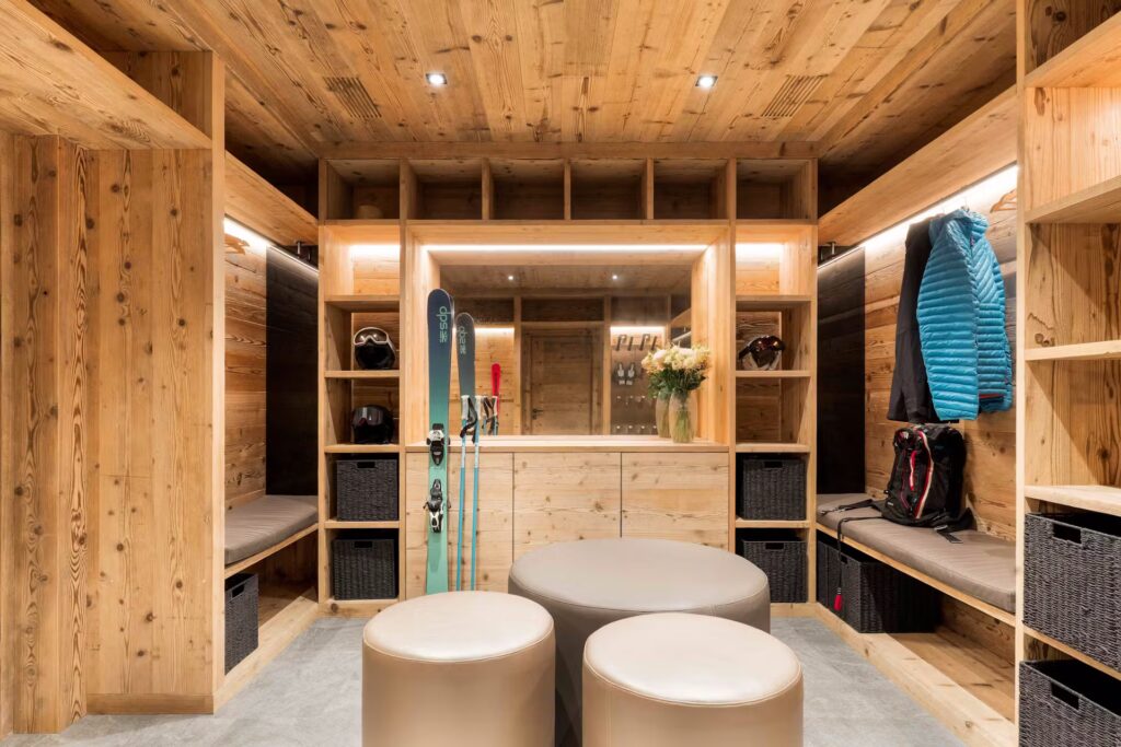 A cozy wooden ski room with built-in shelves, storage baskets, benches, a blue jacket, backpacks, and skis. Two round ottomans and a vase with flowers are in the center under warm ceiling lights. David Pearson Travel