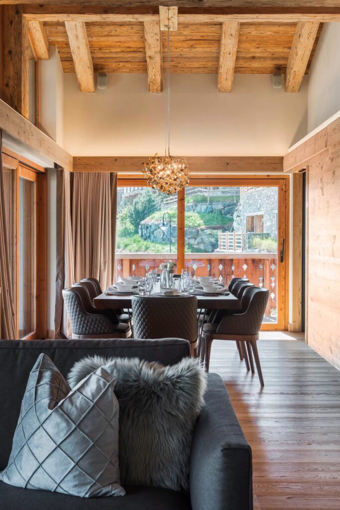 A cozy dining area with a wooden ceiling and walls, large windows overlooking a mountain view, a modern chandelier above a set table, and a sofa with decorative pillows in the foreground. David Pearson Travel