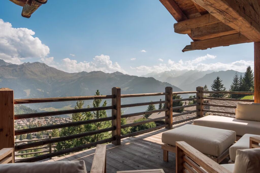 A wooden terrace with cushioned seating overlooks a scenic mountain landscape, with a village below, pine trees, and distant peaks under a clear, sunny sky. David Pearson Travel