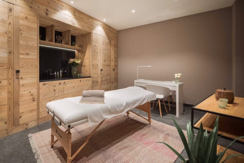 A modern, cozy spa room with a massage table covered in white linens, a towel, and flowers. Wooden cabinets, a desk with a chair, soft lighting, and neutral tones create a serene atmosphere. David Pearson Travel
