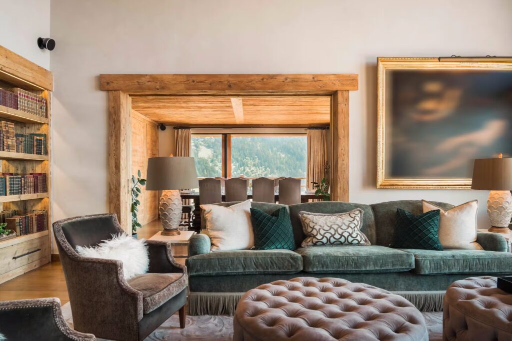 A cozy living room with a green velvet sofa, cushioned chairs, two patterned ottomans, and bookshelves. Large windows in the adjoining dining area reveal a view of green trees and mountains outside. David Pearson Travel