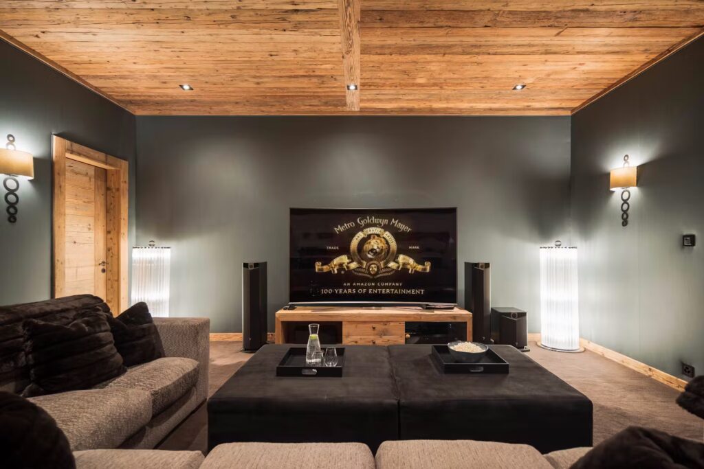 A cozy home theater room with a large flat-screen TV displaying the MGM logo, plush beige sofas, dark ottomans, floor lamps, and wood-paneled ceiling and doors, creating a warm and inviting atmosphere. David Pearson Travel