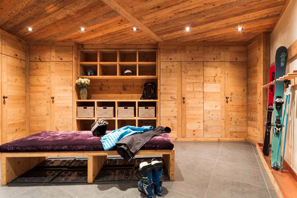 A cozy wooden ski room with built-in cabinets, shelves, and baskets. A bench with winter gear, helmet, boots, and a jacket is in the center. Skis and snowboards are neatly stored against the right wall. David Pearson Travel