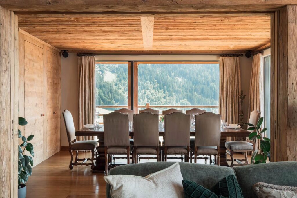 A rustic dining room with a wooden ceiling and large windows overlooking a forested mountain view. A long table with cushioned chairs is set for a meal, and potted plants sit in the corners of the cozy space. David Pearson Travel