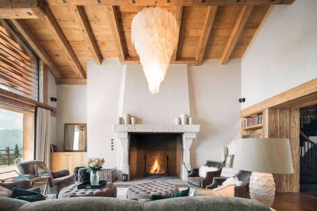 A cozy living room with a high wooden ceiling, large fireplace, elegant chandelier, armchairs, bookshelves, and a large window showing a mountain view. A fire burns in the fireplace, creating a warm, inviting atmosphere. David Pearson Travel