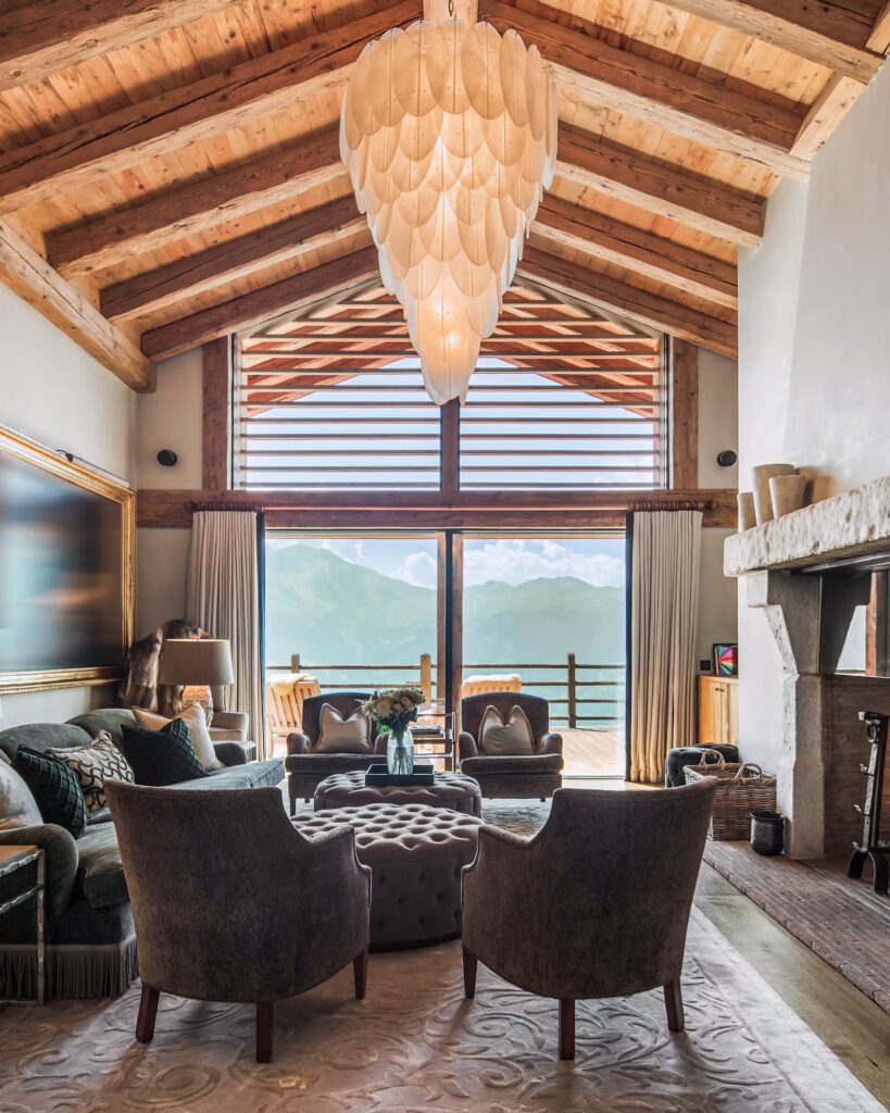 A cozy, elegant living room with plush chairs and a sofa, a large chandelier, a stone fireplace, and floor-to-ceiling windows revealing a mountain view and balcony under a wood-beamed ceiling. David Pearson Travel