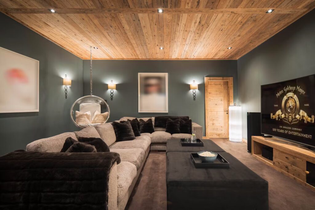A cozy modern living room with a large beige sectional sofa, dark throw pillows, a hanging bubble chair, a wooden ceiling, a wall-mounted TV, dim lighting, and abstract artwork on dark-colored walls. David Pearson Travel