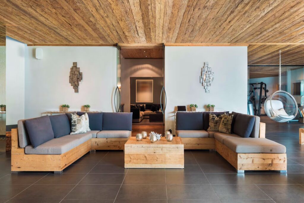 Modern lounge with wooden ceiling and two L-shaped wooden sofas with gray cushions facing a central coffee table, decorative wall art, and plants, with a gym area visible in the background through an open doorway. David Pearson Travel