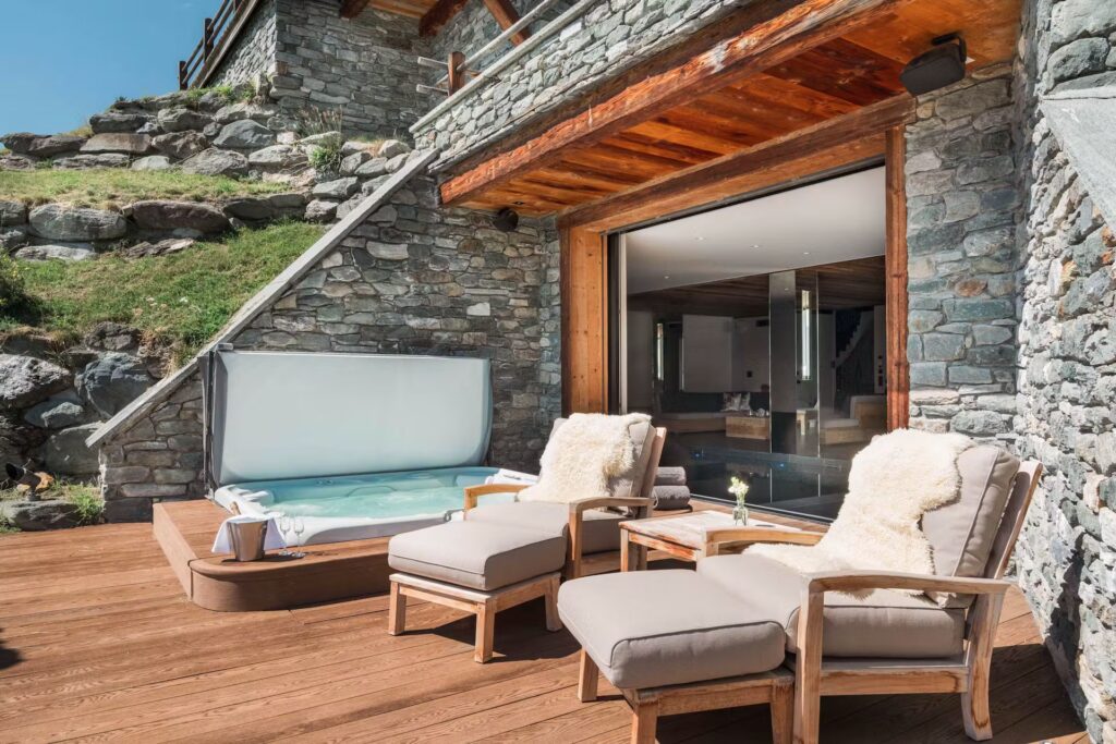 A cozy outdoor patio features two cushioned lounge chairs with fur throws beside a small table and a hot tub, set against a rustic stone building with large glass doors and a wooden deck. David Pearson Travel