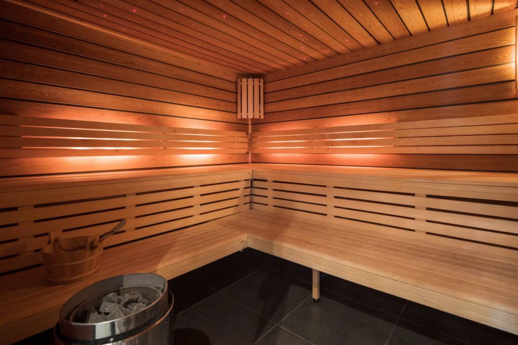 Modern wooden sauna interior with light wooden benches along the walls, soft warm lighting, a bucket with a ladle, and a metal stove with stones on a dark tiled floor. David Pearson Travel