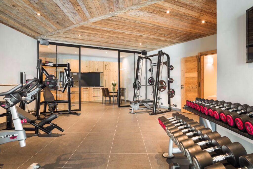 Modern gym room with large windows, wooden ceiling, and tiled floor. Various gym equipment, such as dumbbells, weight machines, and benches, are arranged neatly. A small sitting area is visible in the background. David Pearson Travel