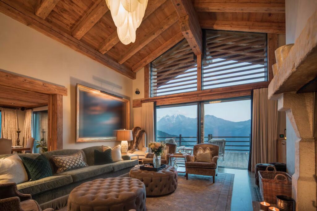 A cozy living room with wooden beams, plush seating, a fireplace, and large glass doors opening to a balcony with mountain views at sunset. Warm lighting and elegant decor create a welcoming atmosphere. David Pearson Travel