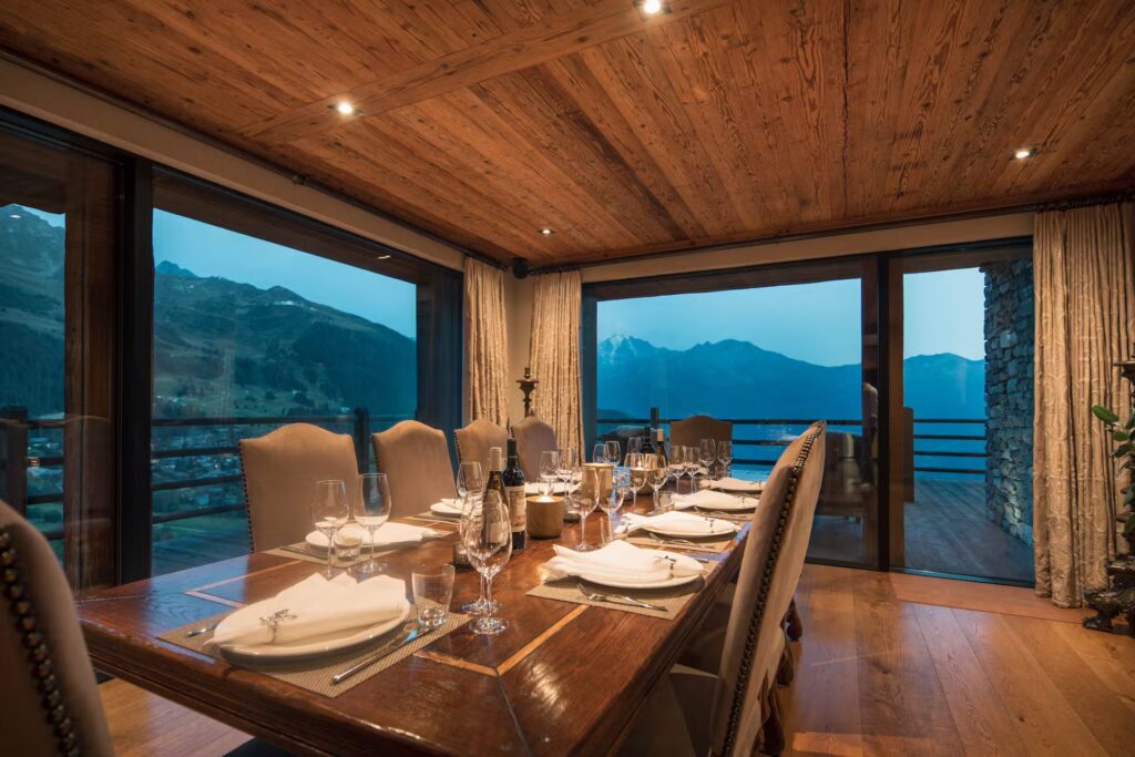 Elegant dining room with a wooden ceiling and large windows offering a scenic view of mountains at dusk; a polished table is set with plates, glasses, and napkins, surrounded by cushioned chairs. David Pearson Travel