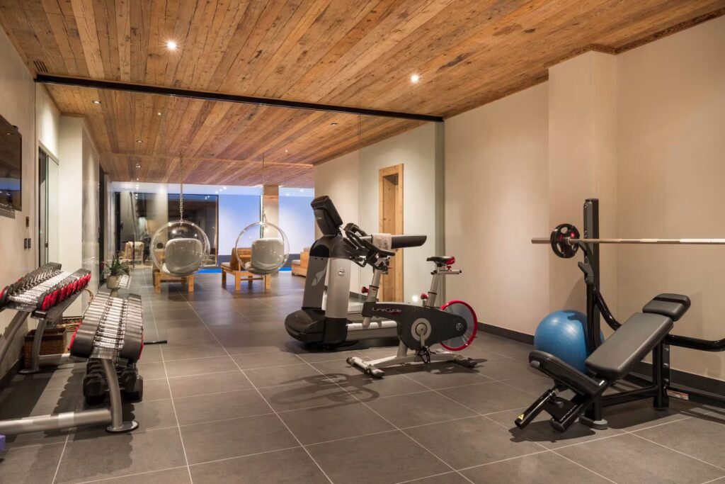 A modern home gym with dumbbells, a treadmill, a stationary bike, a weight bench, barbell, exercise ball, and mirrored wall; lounge area with hanging chairs visible in the background. David Pearson Travel