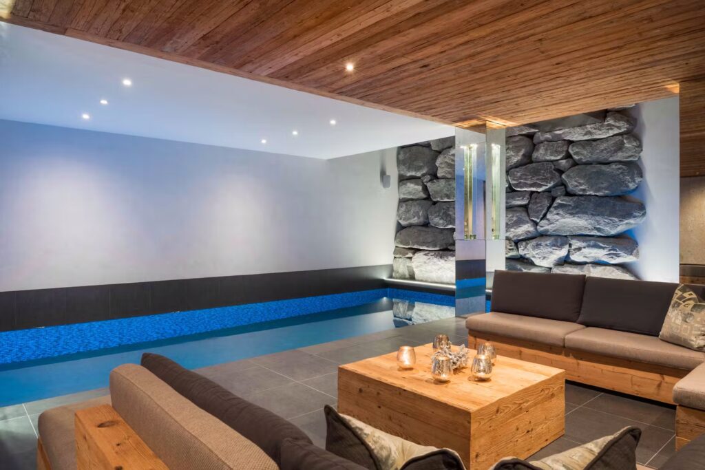 Modern indoor swimming pool with blue water, surrounded by wooden ceiling panels, stone accent walls, and cozy seating areas with wooden furniture and gray cushions. Silver tea sets are placed on the wooden tables. David Pearson Travel
