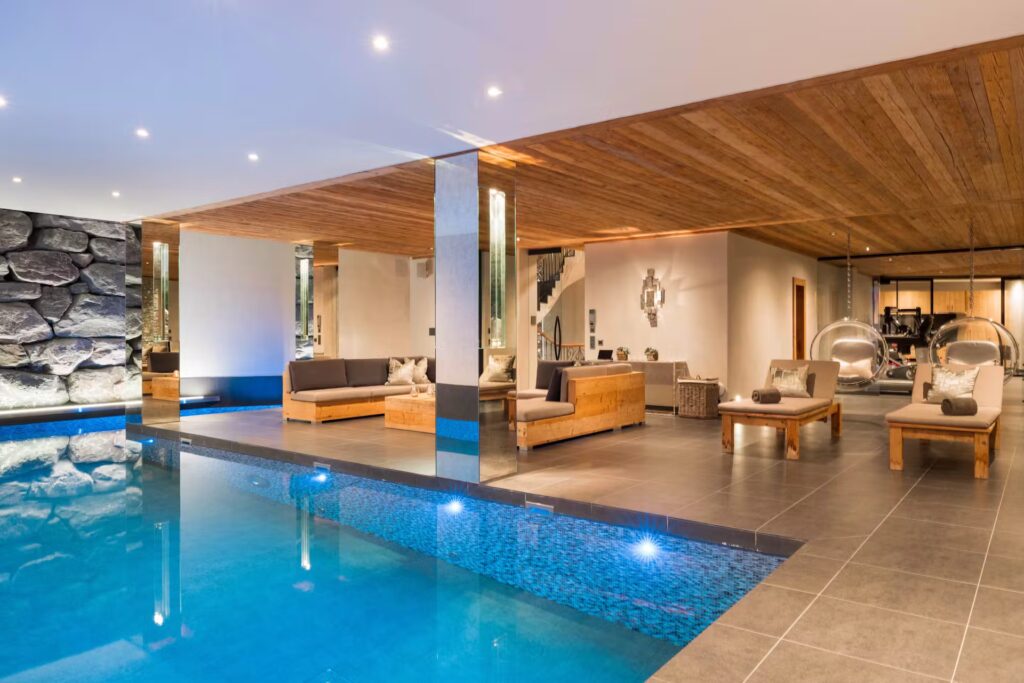 A modern indoor pool area with wooden ceilings, stone walls, and built-in pool lights. The space features wooden lounge furniture, hanging chairs, and a view of a home gym in the background. David Pearson Travel