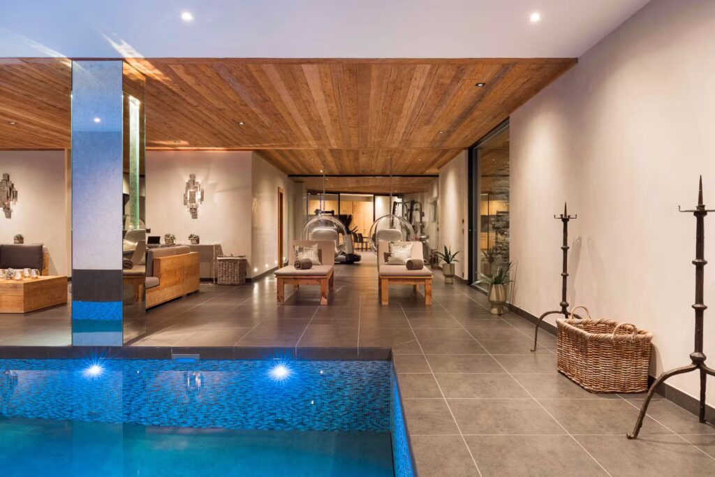 Modern indoor pool area with a wooden ceiling, tiled floor, lounge chairs, hanging bubble chairs, and stylish decor. Warm lighting creates a relaxing, luxurious atmosphere. David Pearson Travel