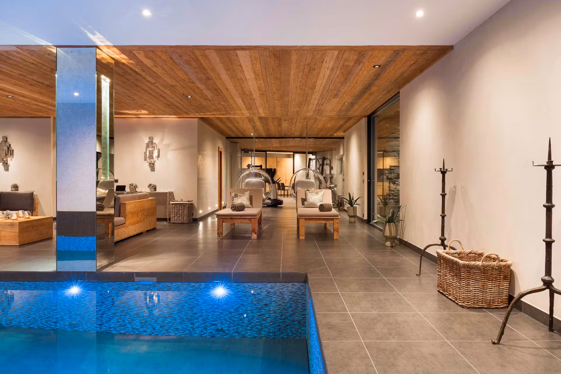 Modern indoor pool area with a wooden ceiling, tiled floor, lounge chairs, hanging bubble chairs, and stylish decor. Warm lighting creates a relaxing, luxurious atmosphere. David Pearson Travel