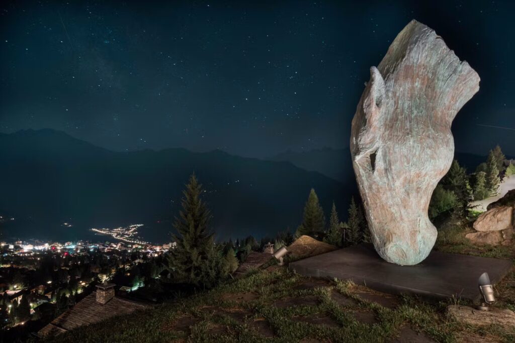 A large horse head sculpture stands on a grassy hill at night, overlooking a town with glowing lights. Pine trees and distant mountains are visible under a starry sky. David Pearson Travel