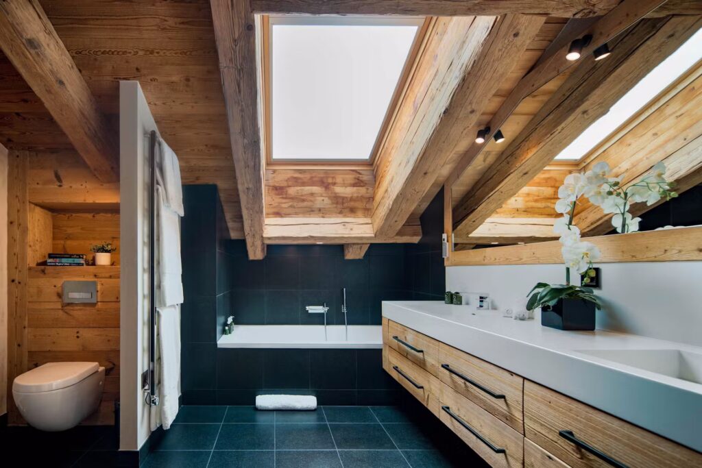 Modern bathroom with wooden beams, large skylights, a black tile floor, freestanding bathtub, double sink with wooden cabinets, white orchid, and wall-mounted toilet. Natural light fills the space. David Pearson Travel