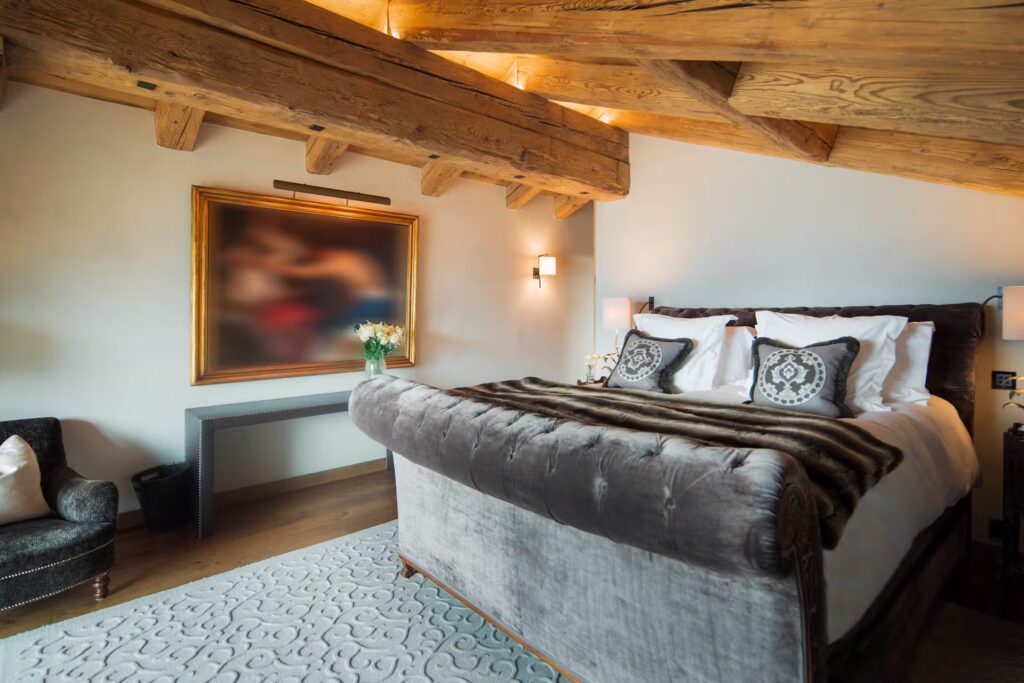 A cozy bedroom with a large velvet bed, patterned pillows, fur throw, decorative carpet, and exposed wooden beams. A blurred framed picture hangs above a console table with flowers; warm lighting creates a welcoming feel. David Pearson Travel