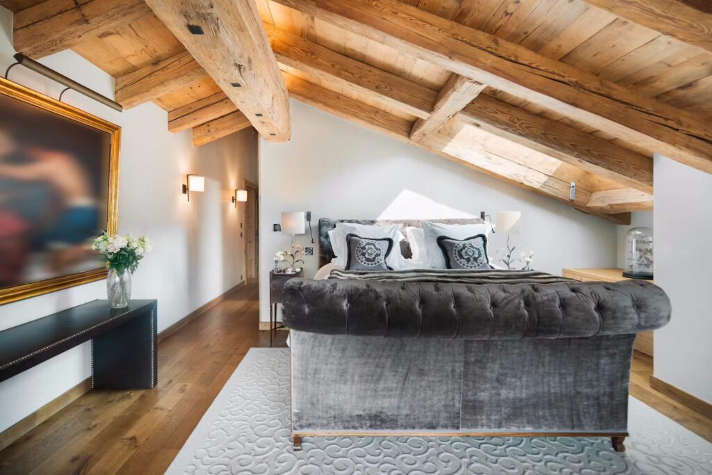 A cozy attic bedroom with exposed wooden beams, a large tufted gray bed, elegant bedding, a white rug, modern wall sconces, fresh flowers on a sleek black console, and a blurred framed artwork on the wall. David Pearson Travel