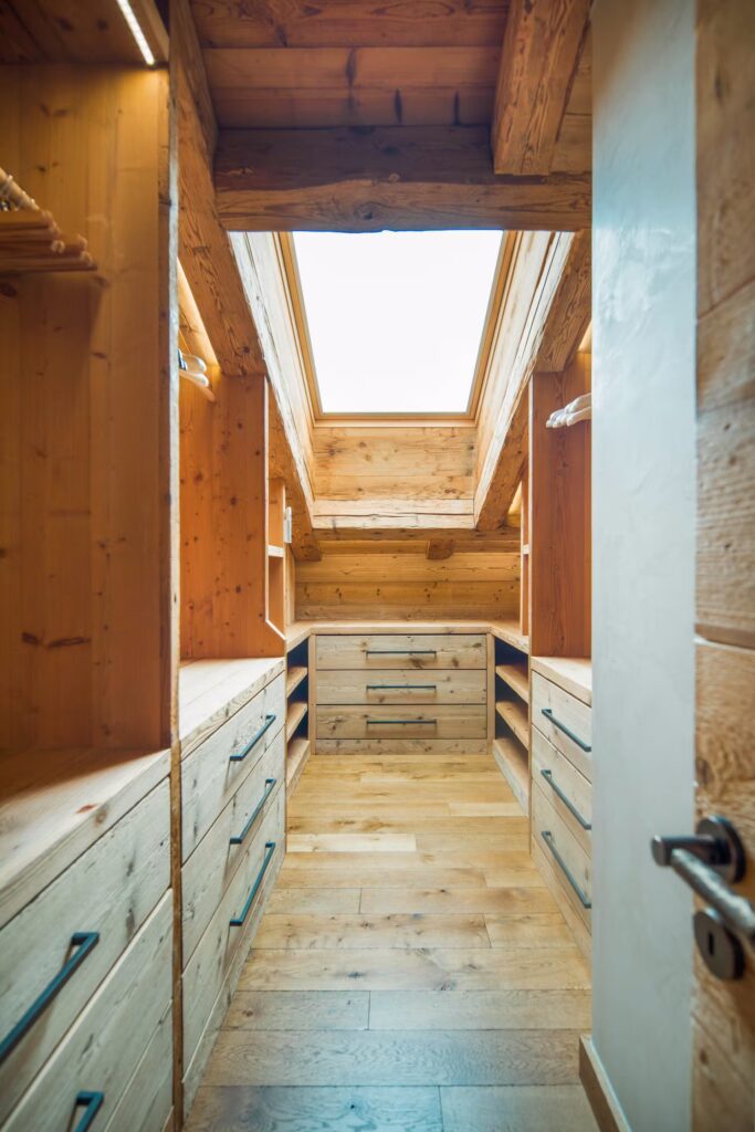 A wooden walk-in closet with drawers and shelves on both sides, illuminated by natural light from a large skylight window in the sloped ceiling above. David Pearson Travel