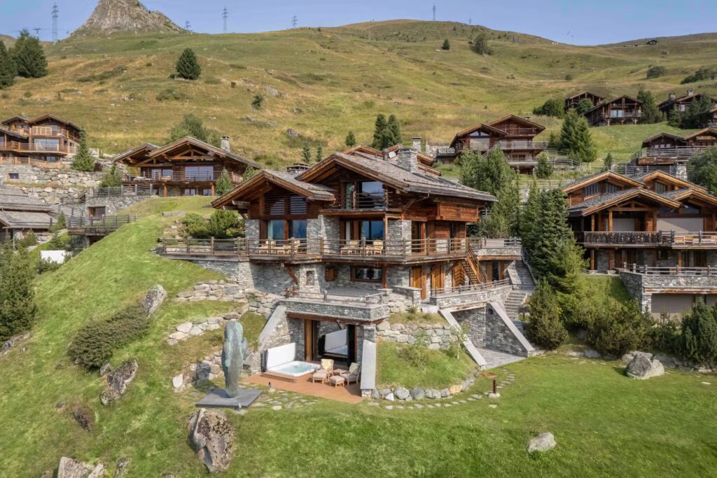 A large wooden chalet with stone accents sits on a green hillside, surrounded by similar alpine houses and rolling grassy slopes under a clear blue sky. A small patio and sculpture are visible in front. David Pearson Travel