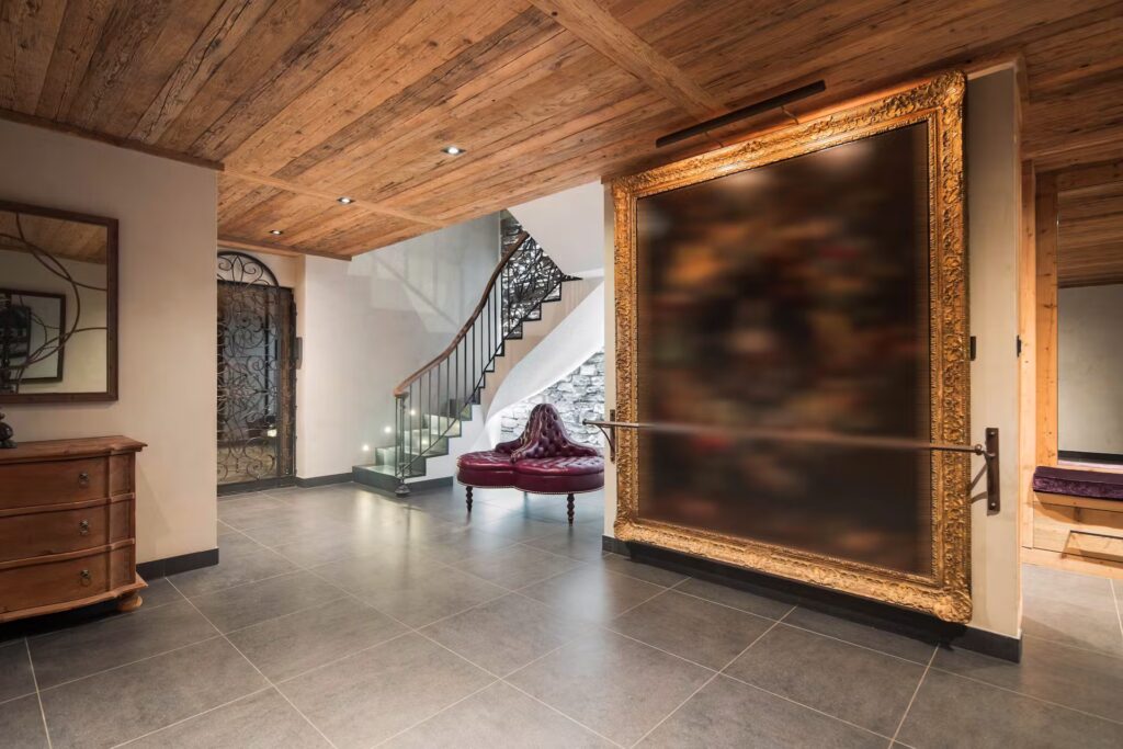 A spacious hallway with a wooden ceiling, tiled floor, a staircase with a wrought iron railing, a large ornate framed artwork on the wall, and a red tufted bench beneath a draped fabric. David Pearson Travel