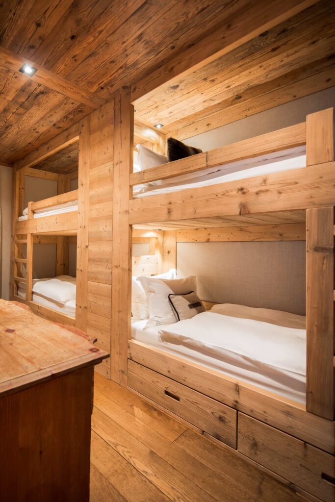 A cozy wooden room with two sets of bunk beds, white bedding, and built-in drawers beneath the lower bunks. The walls, ceiling, and furniture are made of light-colored wood, creating a warm, rustic atmosphere. David Pearson Travel