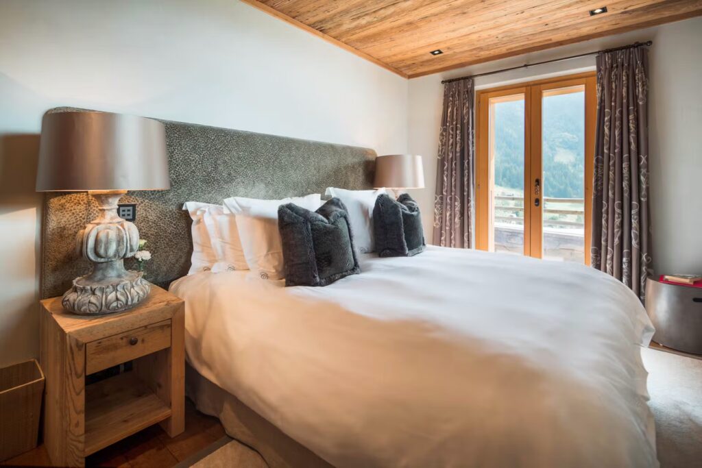 A cozy bedroom with a large bed, white bedding, two dark throw pillows, wooden nightstands with lamps, and glass doors leading to a balcony with a mountain view. The ceiling and accents are made of wood. David Pearson Travel