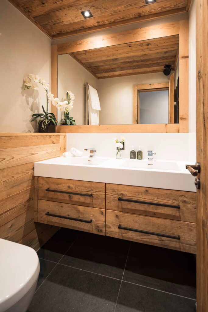 A modern bathroom with wooden cabinets, a large mirror, double sinks, black drawer handles, white countertop, and orchid flowers in a vase. The room features wood-paneled walls and ceiling, with dark tile flooring. David Pearson Travel